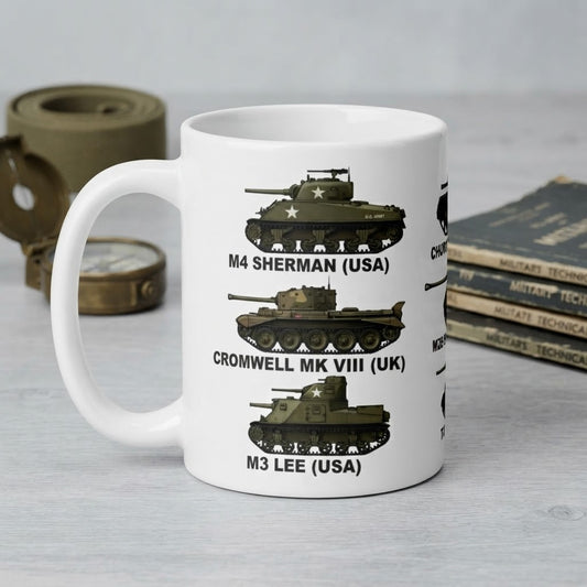 A white ceramic mug printed with military tank illustrations and text reading 'M4 SHERMAN (USA)', 'CROMWELL MK VIII (UK)', and 'M3 LEE (USA)'. The mug is photographed on a light gray tabletop with rolled canvas, a compass, and books in the background.
