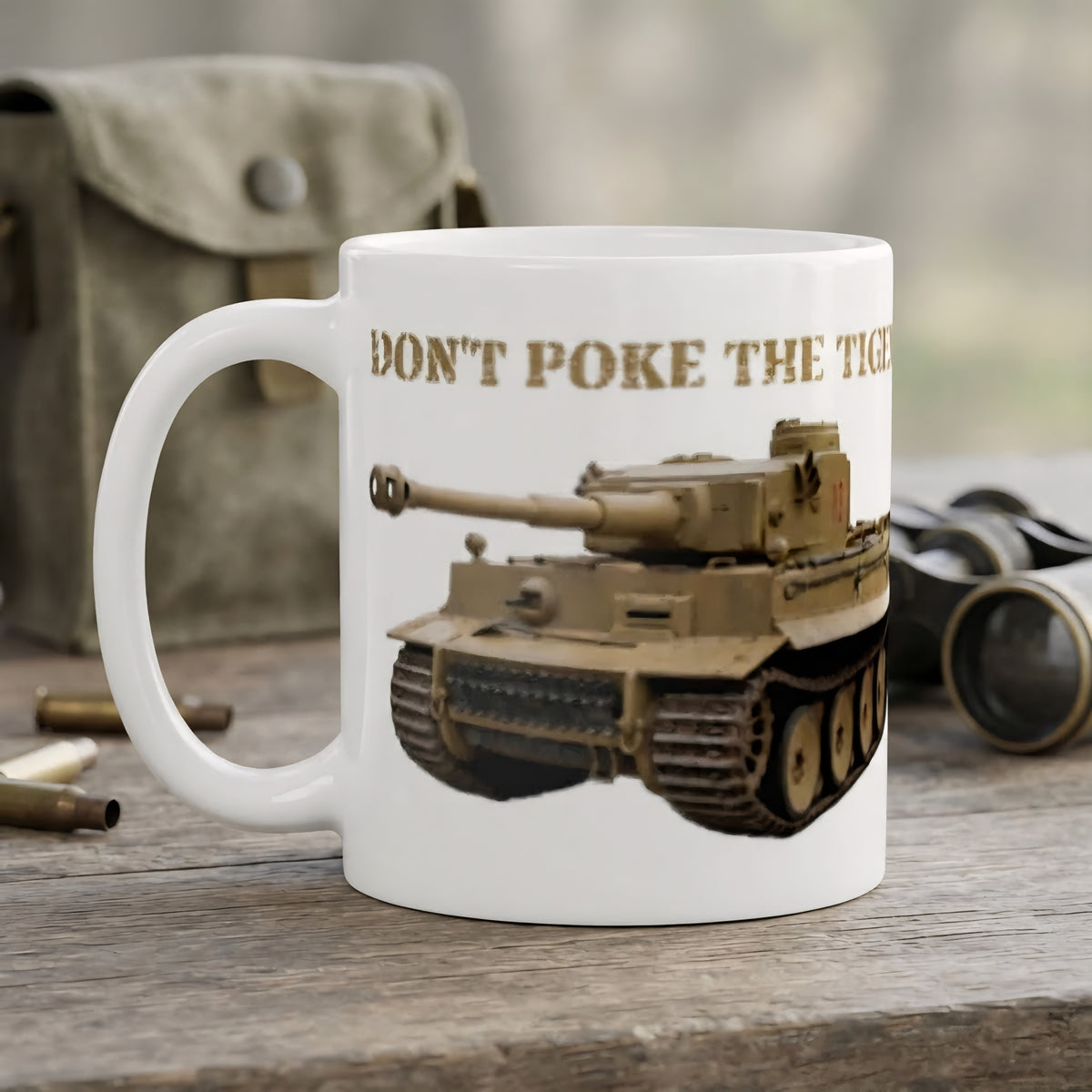 A white ceramic coffee mug printed with a tan military tank illustration and the text 'DON'T POKE THE TIGER' in brown, shown on a rustic wooden surface with blurred vintage props in the background.