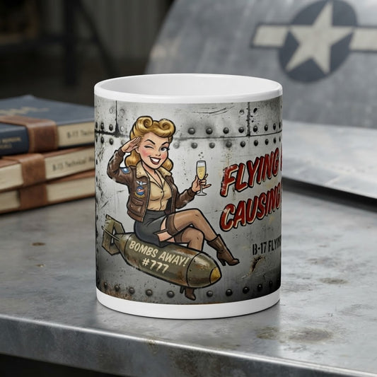 A white ceramic mug with a vintage pin-up bomber illustration sitting on a gray metal tabletop. The mug features a graphic of a blonde woman in a leather jacket and skirt sitting on a bomb with the text 'FLYING CAUSING BOMBS AWAY! #777'.