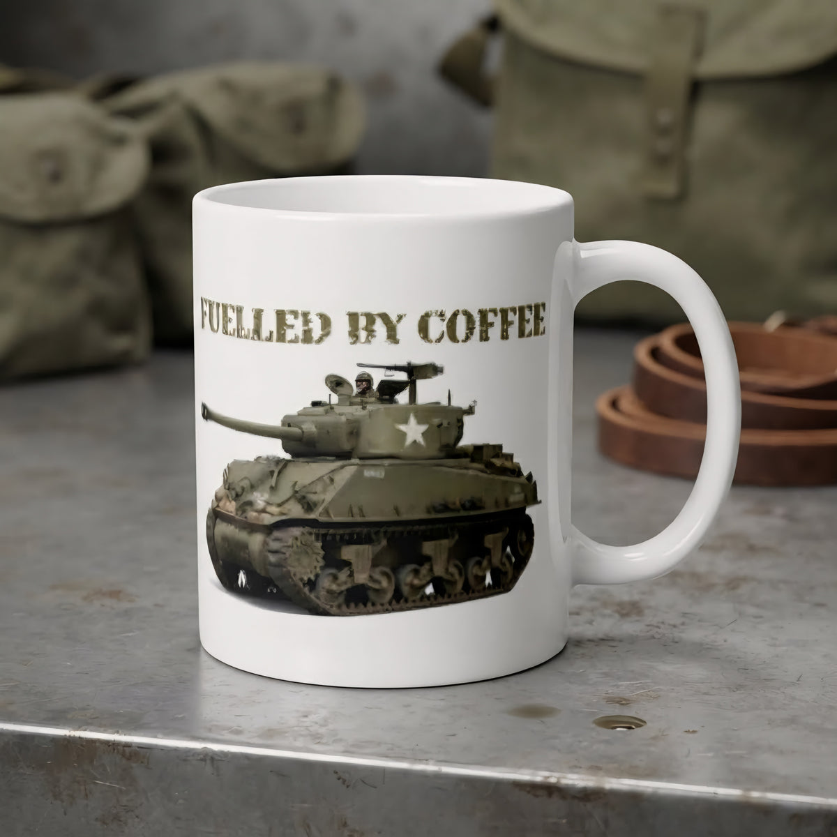 A white ceramic coffee mug with a printed graphic of a military tank and the text 'FUELLED BY COFFEE' in olive drab. The mug is set on a rustic gray metal surface with vintage military-style canvas bags in the blurred background.
