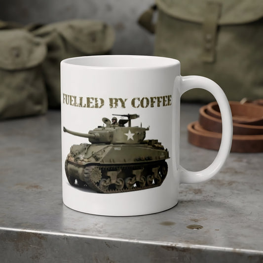 A white ceramic coffee mug with a printed graphic of a military tank and the text 'FUELLED BY COFFEE' in olive drab. The mug is set on a rustic gray metal surface with vintage military-style canvas bags in the blurred background.