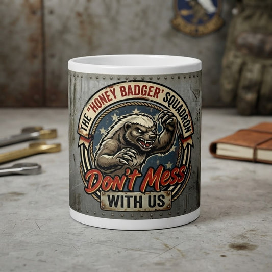 A white ceramic mug with a detailed graphic print of a honey badger and the text 'The "Honey Badger" Squadron Don't Mess With Us', displayed on a rustic, weathered gray tabletop with military-themed props in the background.